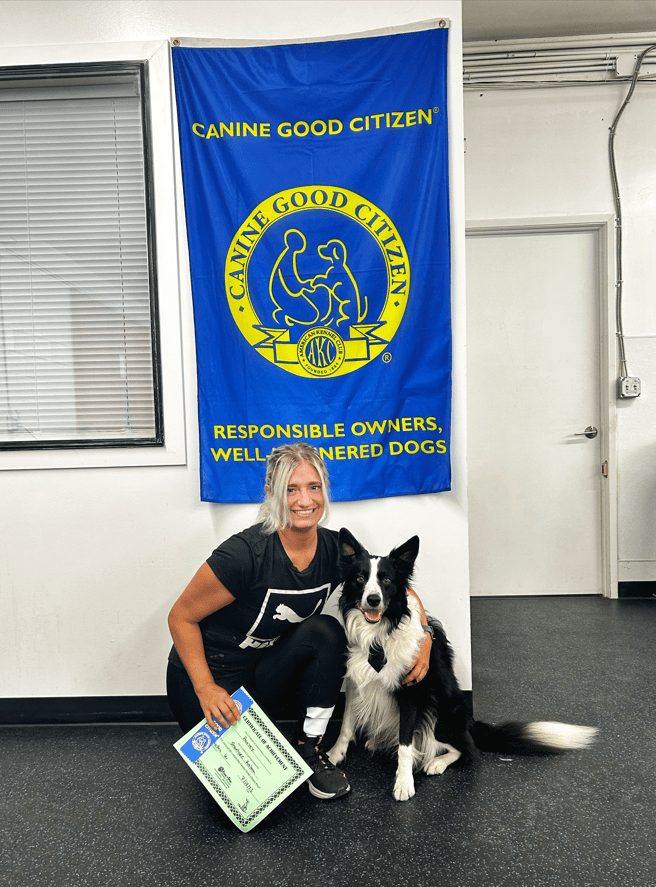 A smiling handler kneeling with her Border Collie and CGC certificate under the Canine Good Citizen banner