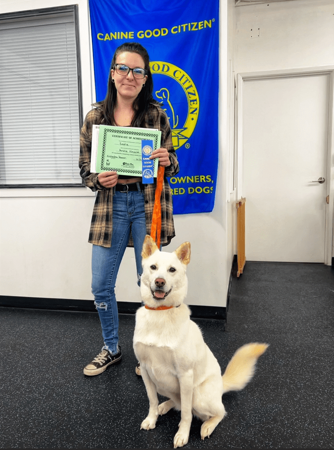 A handler holding a CGC certificate and ribbon, with her white dog sitting in front of the Canine Good Citizen banner