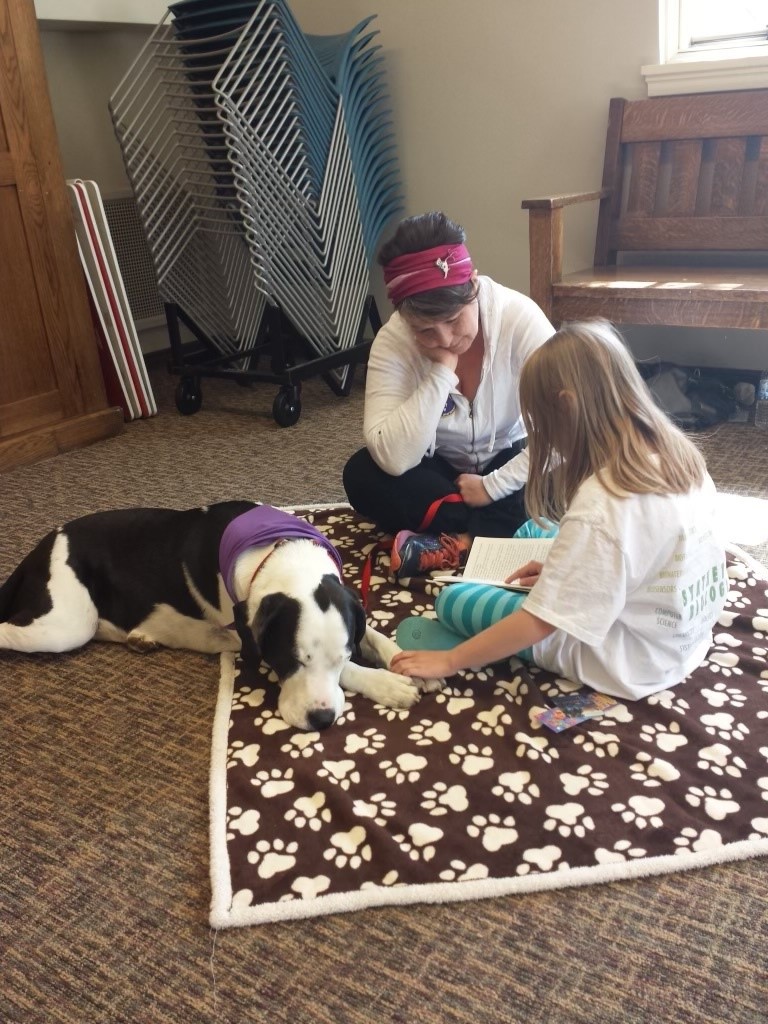A girl reading a book to a black and white therapy dog lying on a paw-print blanket