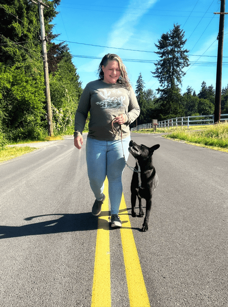 A smiling handler walking her black dog on a loose leash along a country road, the dog looking up at her